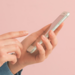 Dłonie w dżinsowej koszuli trzymające smartfon na różowym tle – zdjęcie ilustrujące scrollowanie social mediów i proces rezygnacji z Facebooka oraz Instagrama.