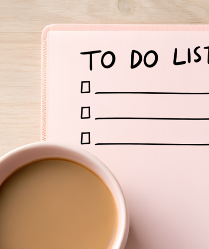 Różowy notatnik z napisem TO DO LIST i puste linie do wypełnienia leżące obok filiżanki herbaty z mlekiem na jasnym drewnianym stole.